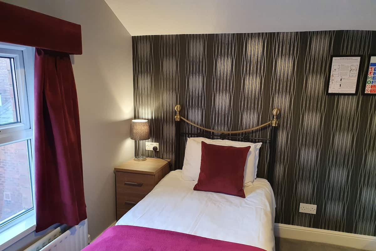 A hotel room with a bed, a nightstand, a lamp, a window with curtains, a radiator, and a wall with a patterned wallpaper.