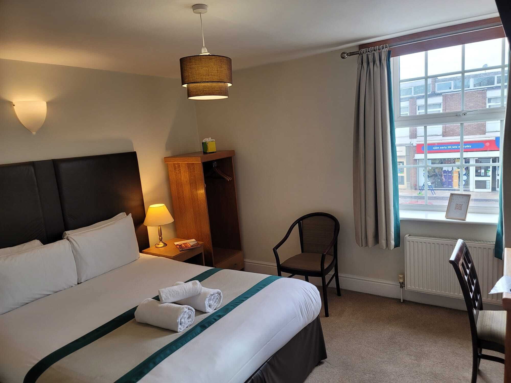 A hotel room with a bed, a chair, a nightstand with a lamp, a window with curtains, and a view of the street outside.