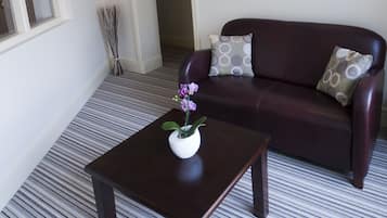 Een woonkamer met een donkerbruine leren bank, een klein houten bijzettafeltje met een vaas paarse bloemen, en een gestreept tapijt.