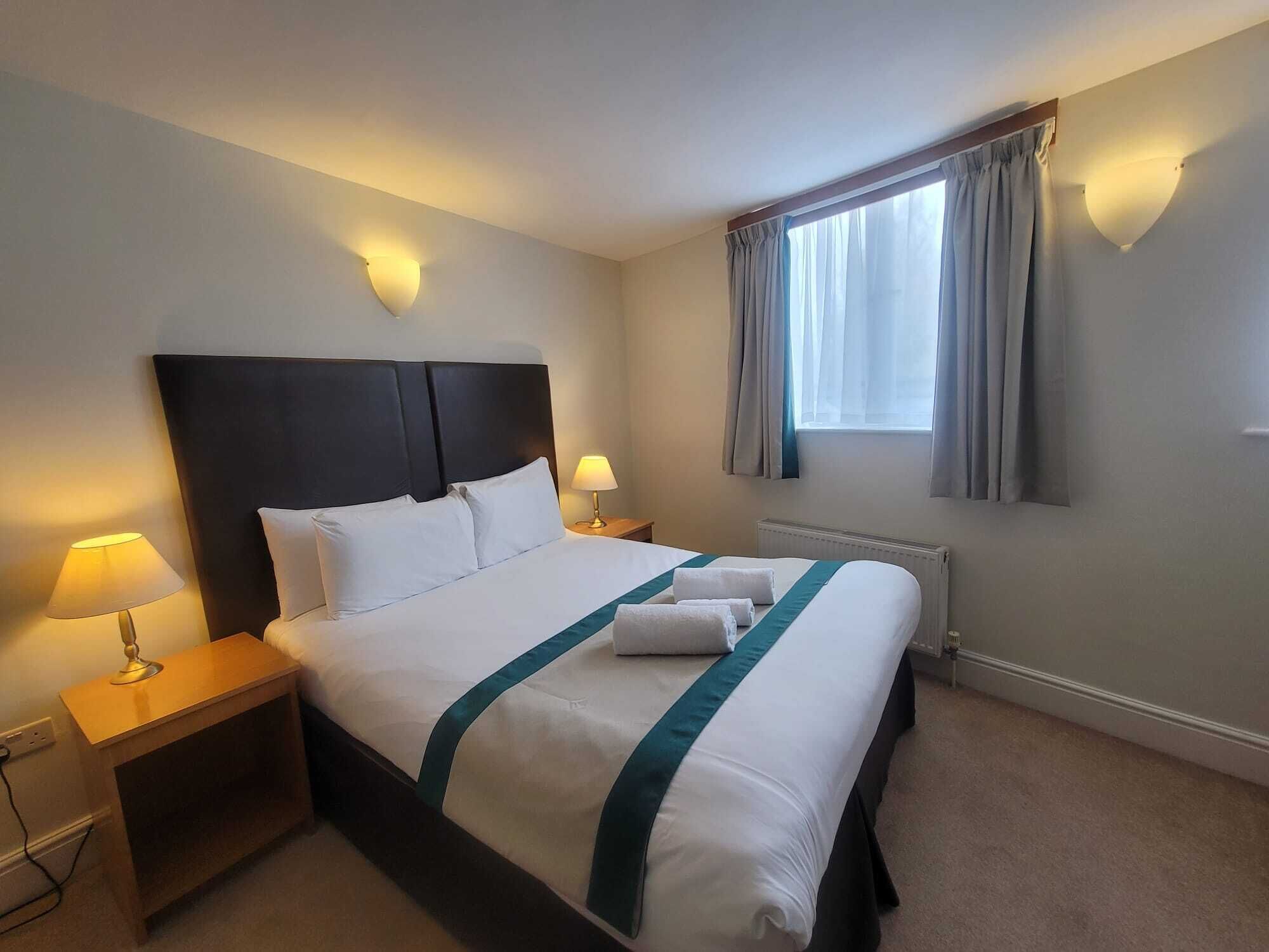 Una habitación de hotel con una cama grande, dos lámparas de noche, una ventana con cortinas y un radiador.