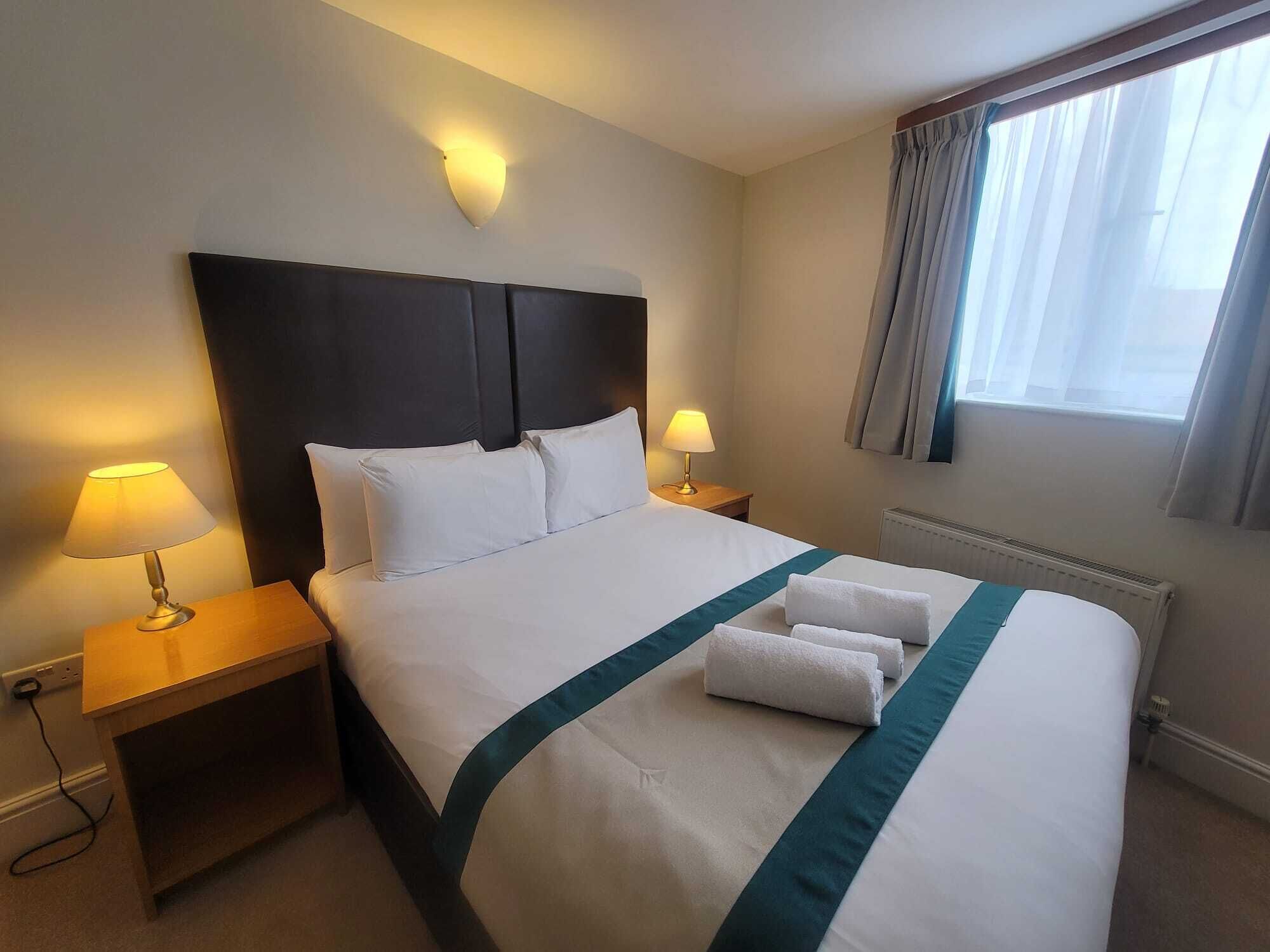 A hotel room with a large bed, two bedside lamps, a headboard, a window with curtains, and a radiator.