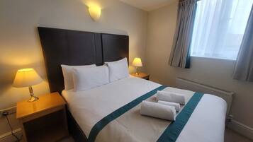 A hotel room with a large bed, two bedside lamps, a headboard, a window with curtains, and a radiator.
