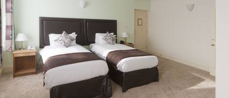 A hotel room with two beds, each with a brown headboard and white bedding. There are bedside tables with lamps and a telephone. The room has a light green wall and a door in the background.