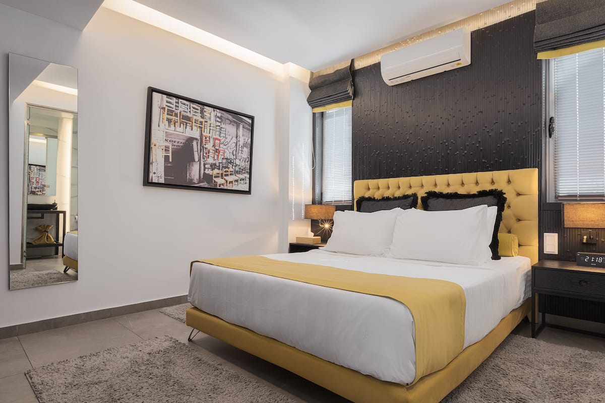 A modern bedroom with a large bed, a bedside table, a nightstand, a wall-mounted air conditioner, and a framed picture on the wall.