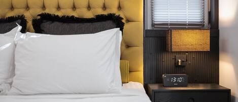 A neatly made bed with white and black pillows, a yellow tufted headboard, a bedside table with a digital clock, and a window with blinds.