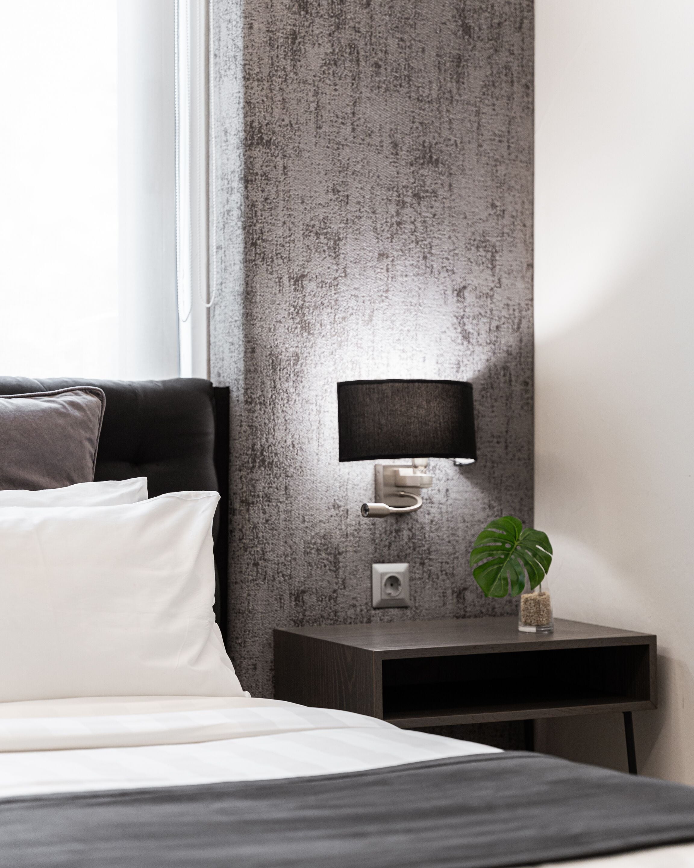 A modern hotel room with a bed, a nightstand with a lamp, and a small plant.