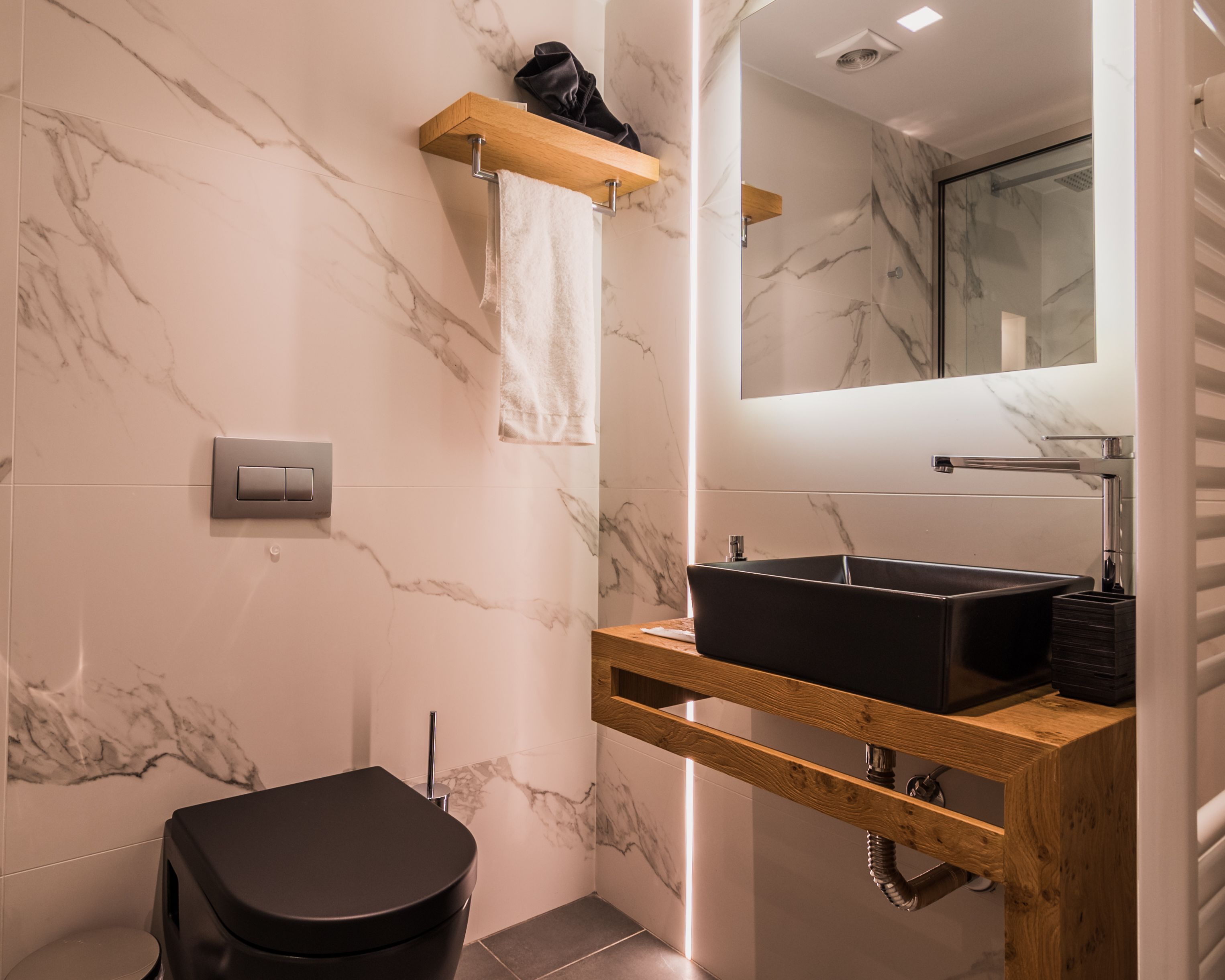 Een moderne badkamer met een marmeren wand, een houten wastafelmeubel, een zwarte wastafel en een toilet.