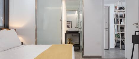 Un dormitorio moderno con una cama, una mesita de noche, un armario y un baño visible a través de una puerta de cristal.