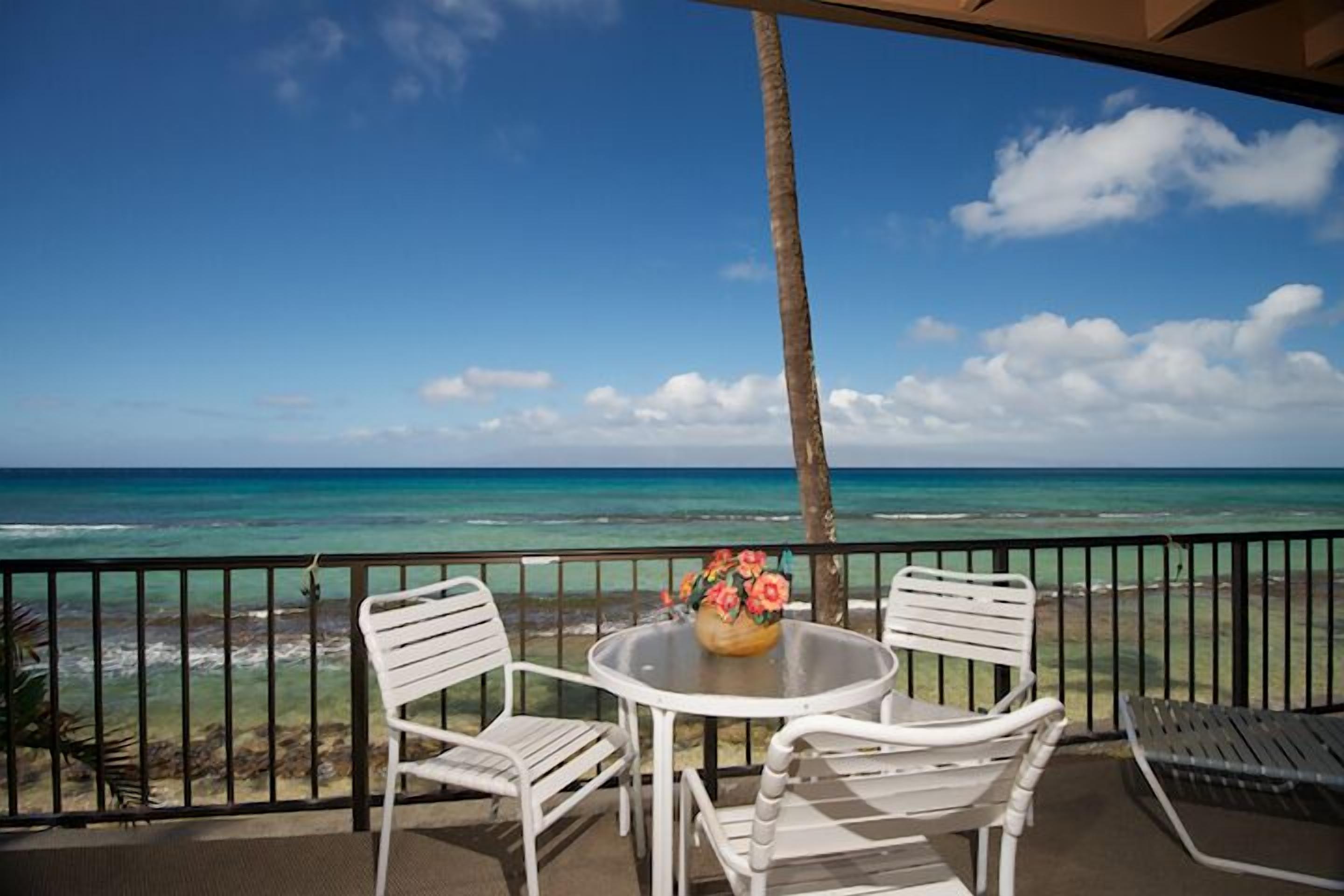 A balcony with white chairs and a table, overlooking a beach and ocean.