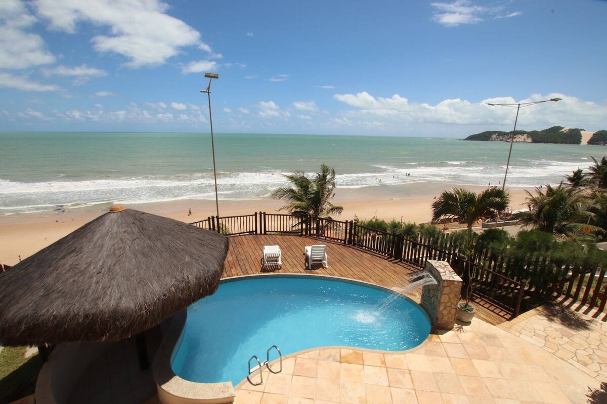 A pool area with a thatched roof, a view of the beach, and a stone deck.