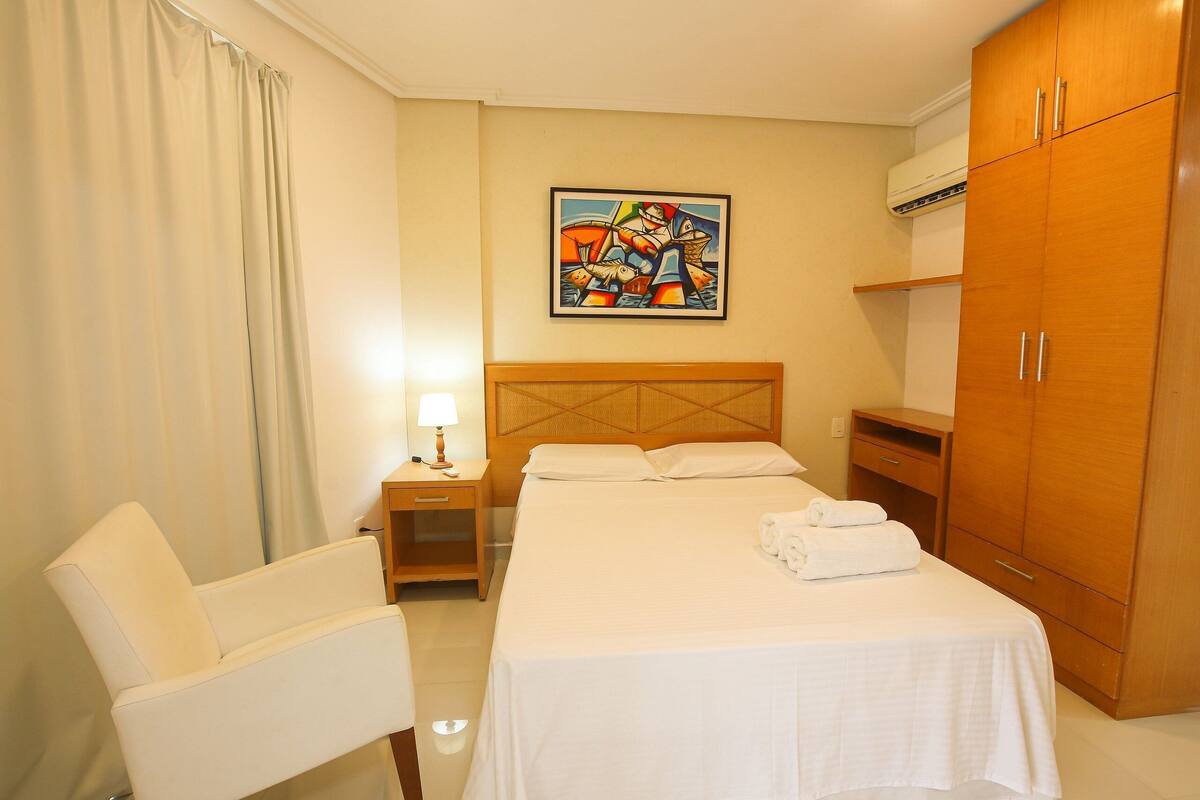 A hotel room with a bed, a chair, a nightstand, a painting on the wall, and a wardrobe.