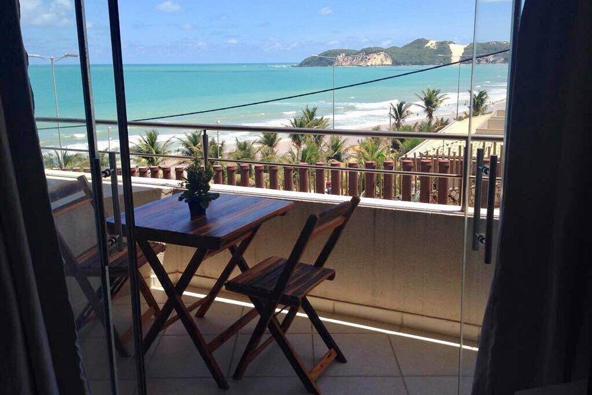 A balcony with a wooden table and chairs, overlooking a beach with clear blue water and a distant mountain.