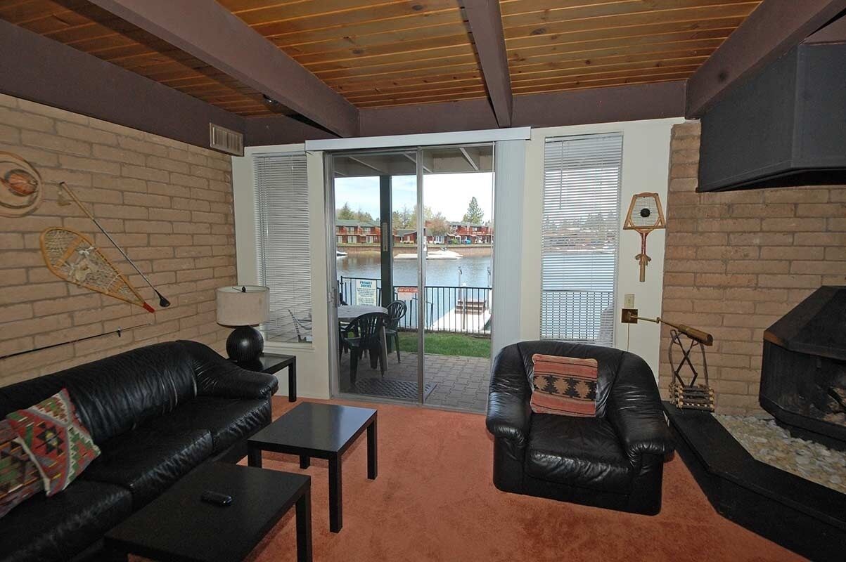 Ein Wohnzimmer mit Kamin, einer schwarzen Ledersofa, einem kleinen Tisch und Blick durch Glastüren auf einen Außenbereich mit Pool und Gebäuden.