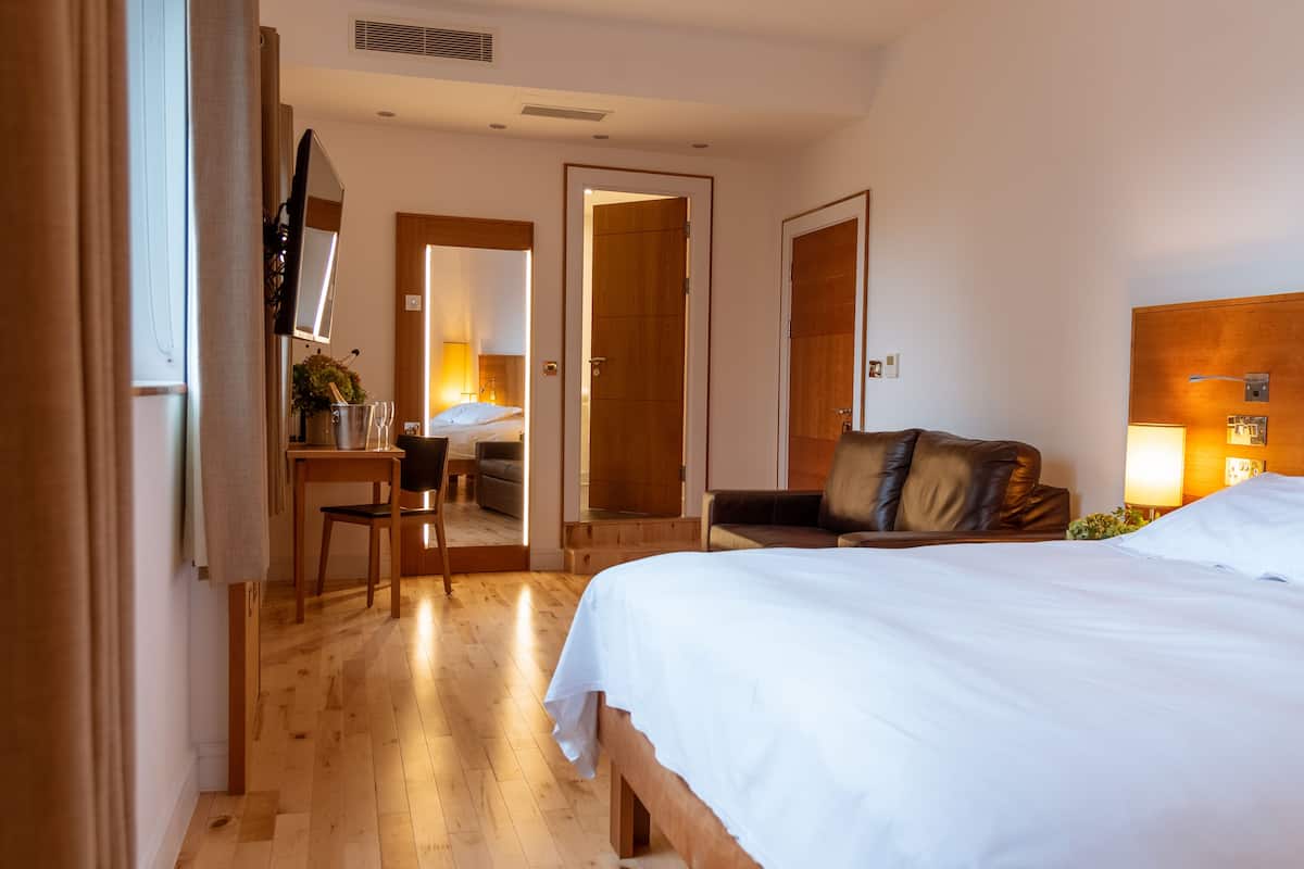 A hotel room with a large bed, a desk with a chair, a wooden floor, and a door leading to another room.