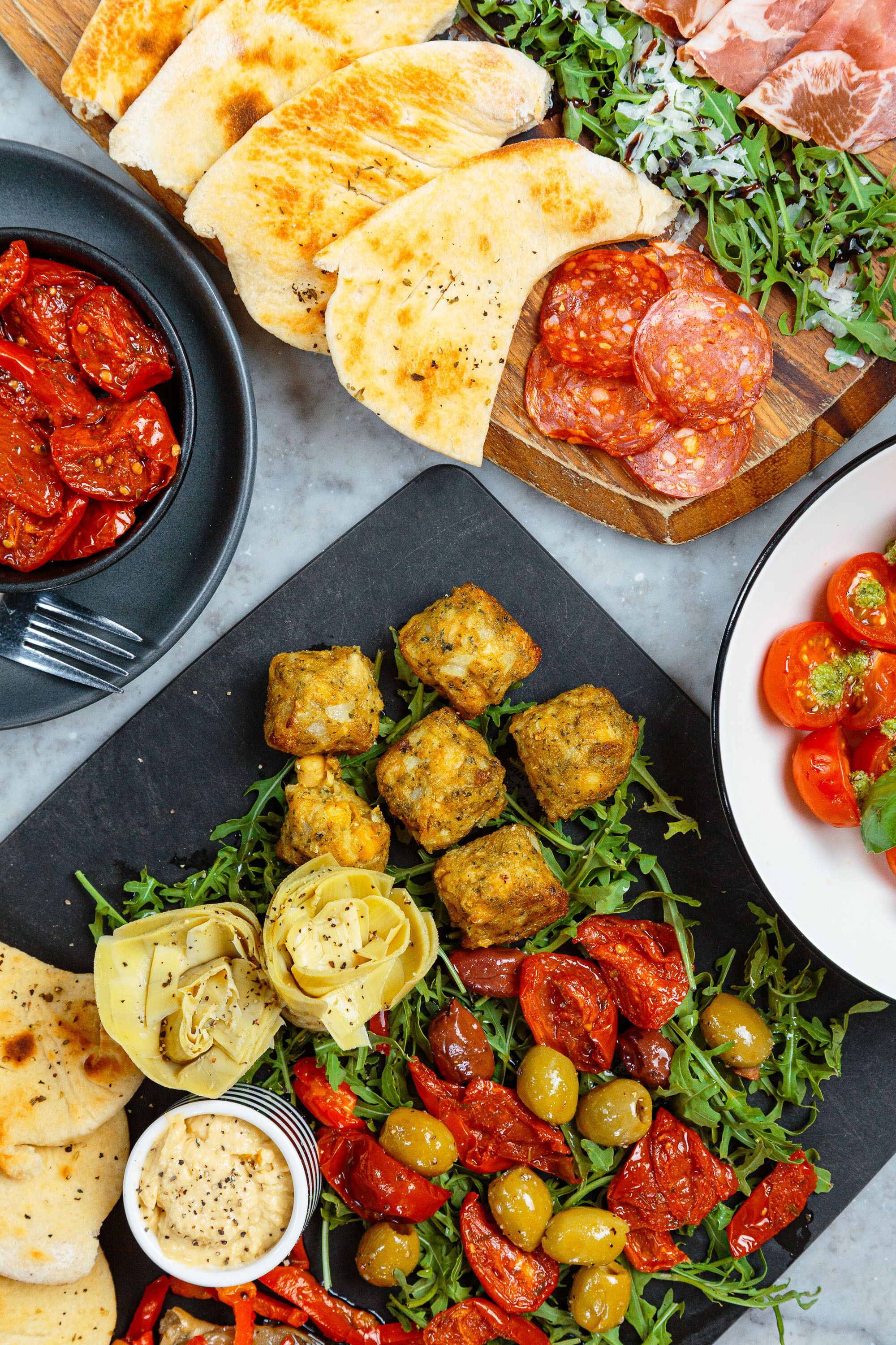 Diversos alimentos como pan plano, salami en rodajas, verduras a la parrilla y un tazón de hummus.
