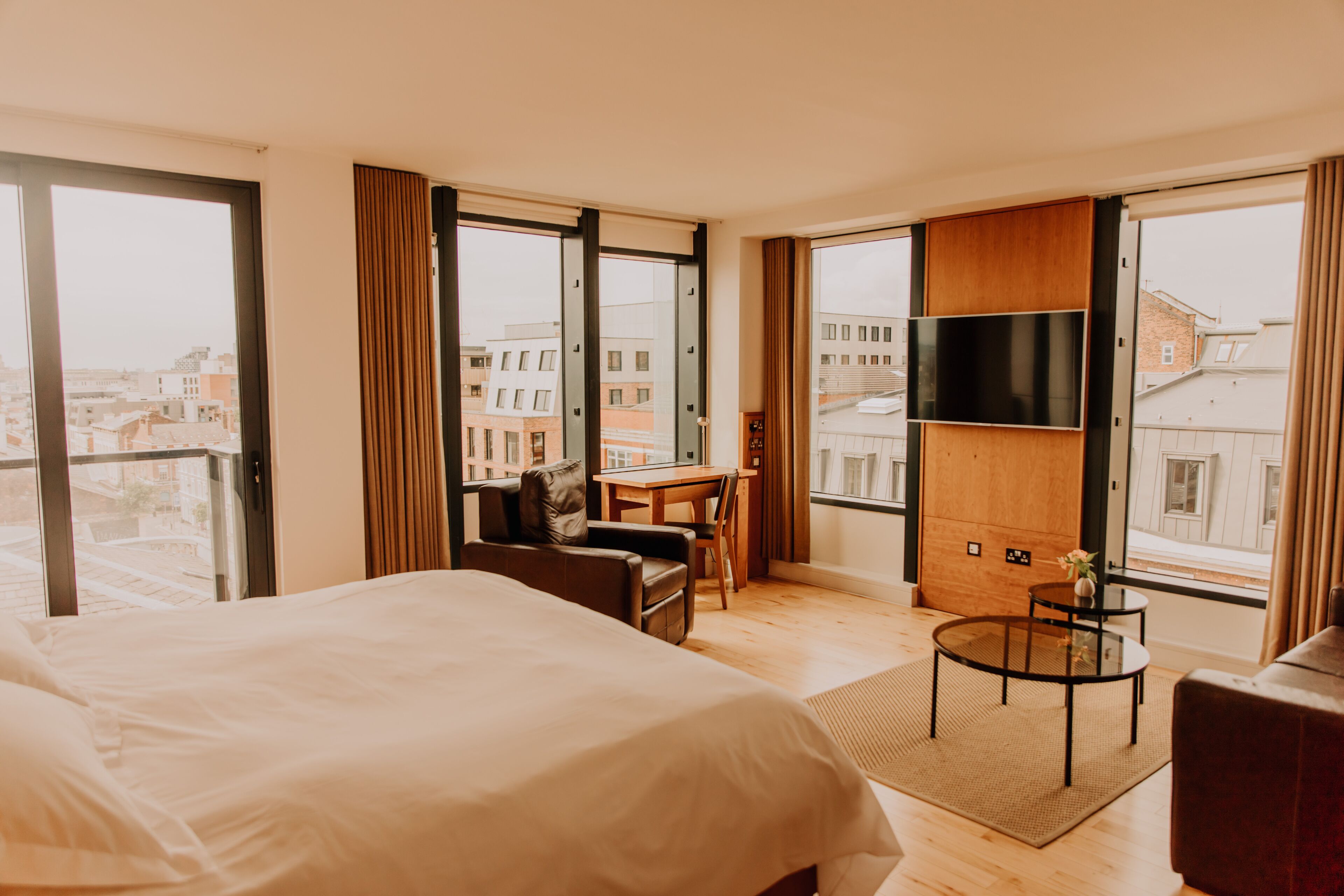 Habitación de hotel con una cama grande, un escritorio, una silla, un televisor y vistas a la ciudad a través de amplios ventanales.