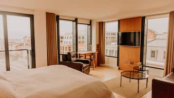 Habitación de hotel con una cama grande, un escritorio, una silla, un televisor y vistas a la ciudad a través de amplios ventanales.