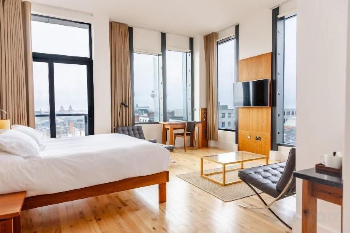 Une chambre d’hôtel équipée d’un lit, d’un bureau, d’une chaise, d’une télévision et de grandes fenêtres donnant sur la ville.