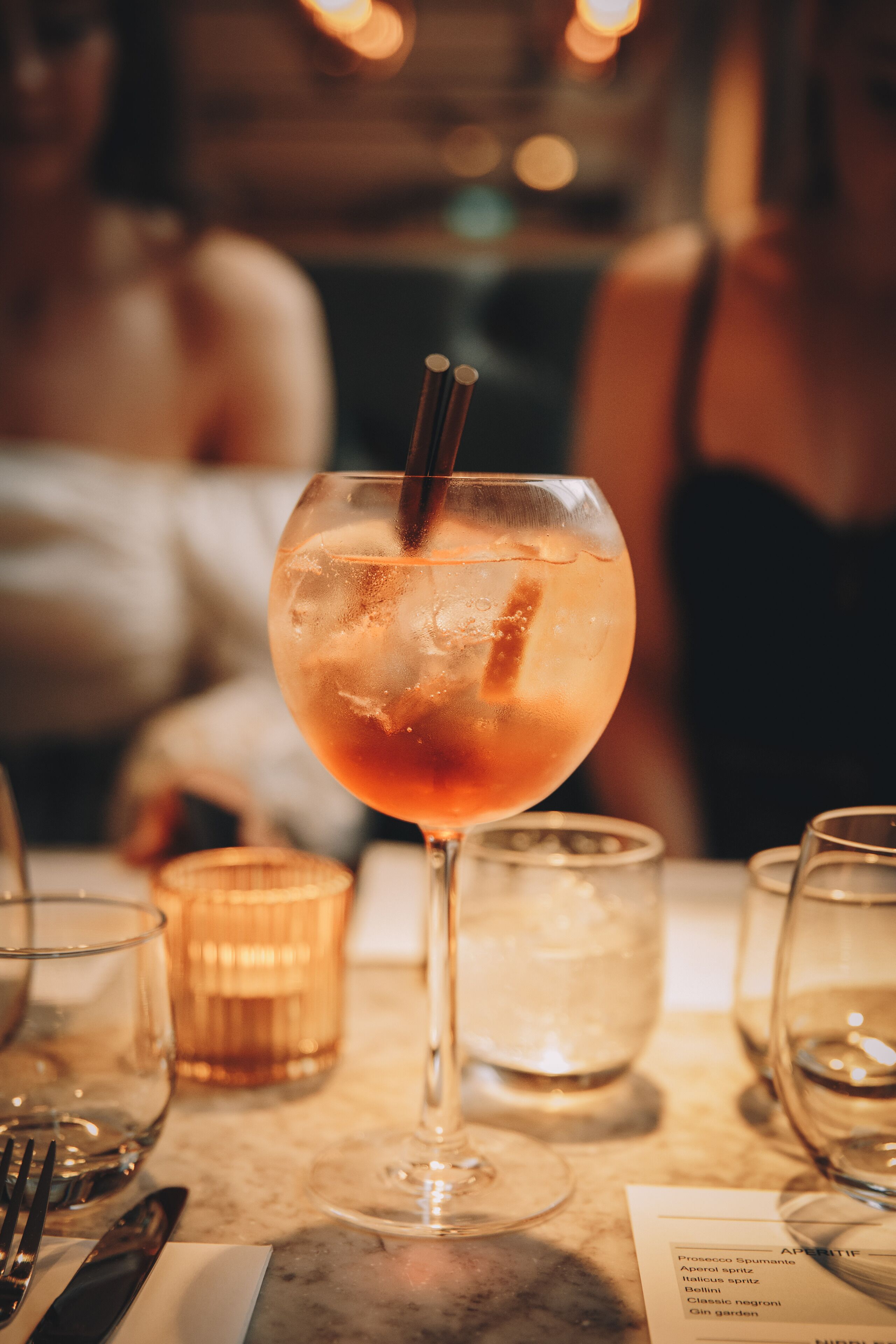 A cocktail glass with a straw on a marble table, surrounded by other glasses and a menu.