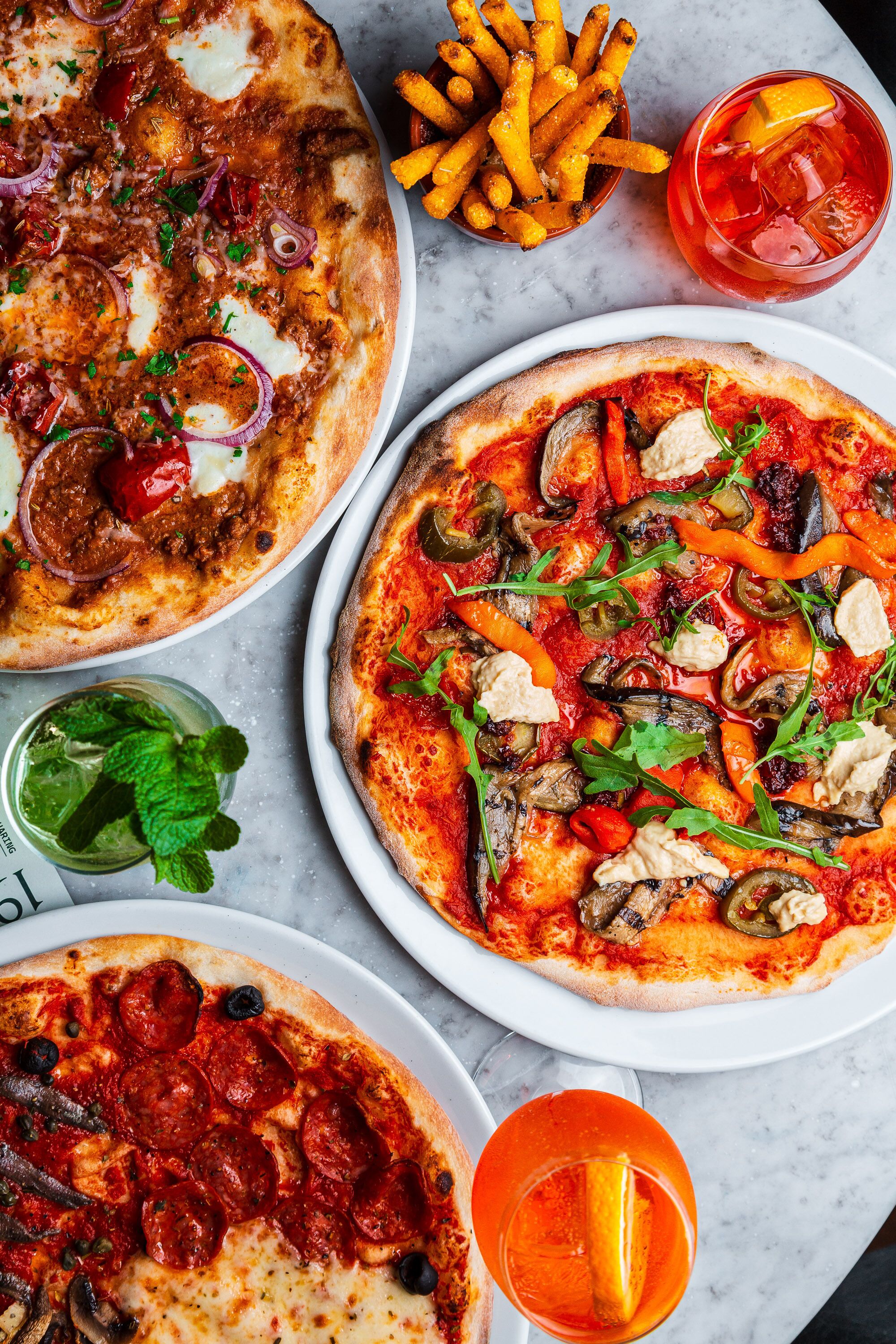 Tres pizzas con diferentes ingredientes, un bol de papas fritas y un vaso de bebida roja sobre una superficie de mármol.