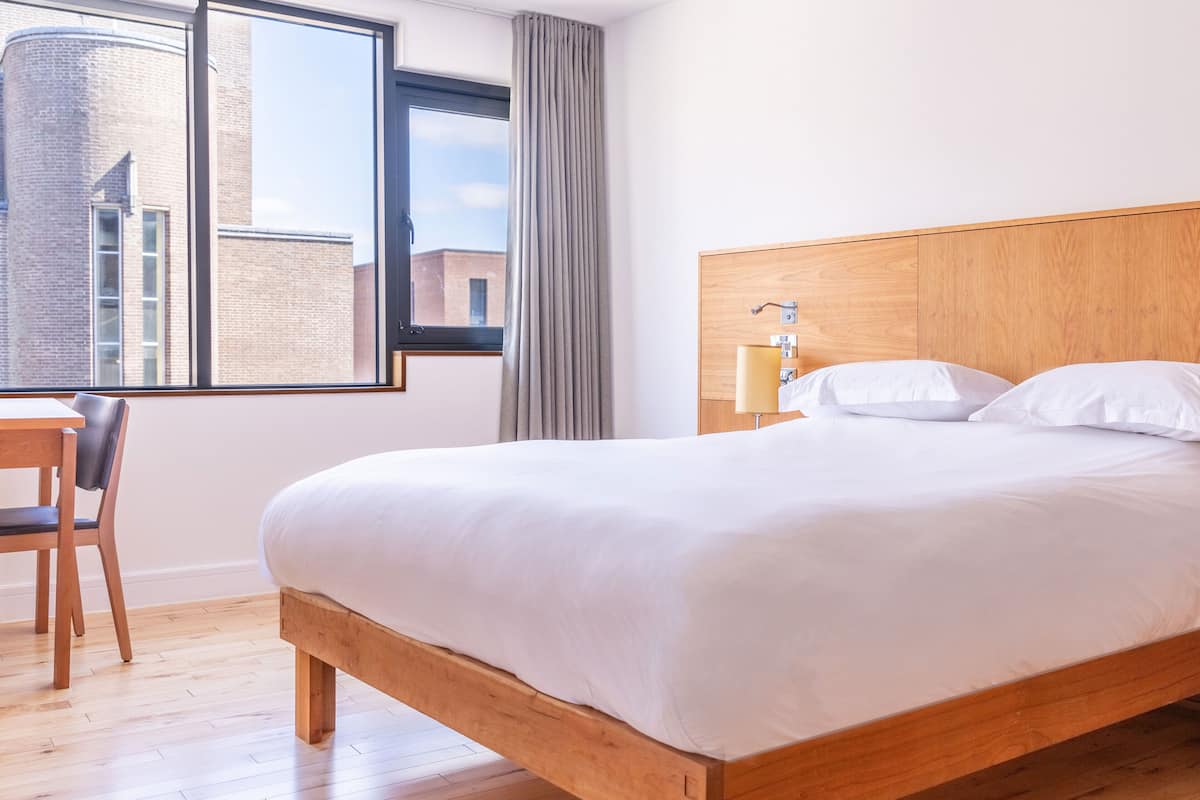 A hotel room with a wooden bed, a desk, and a window with a view of buildings.