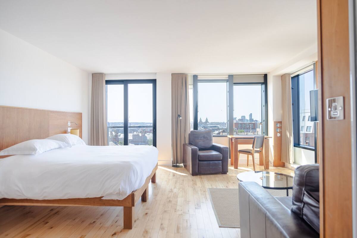 A modern hotel room with a large bed, a seating area with a table, and a view of the city through large windows.