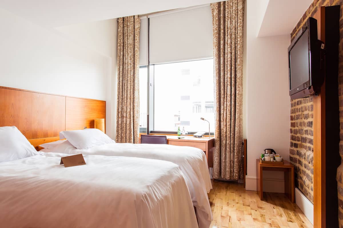 A hotel room with two beds, a wooden headboard, a desk with a chair, a TV mounted on the wall, and a window with curtains.
