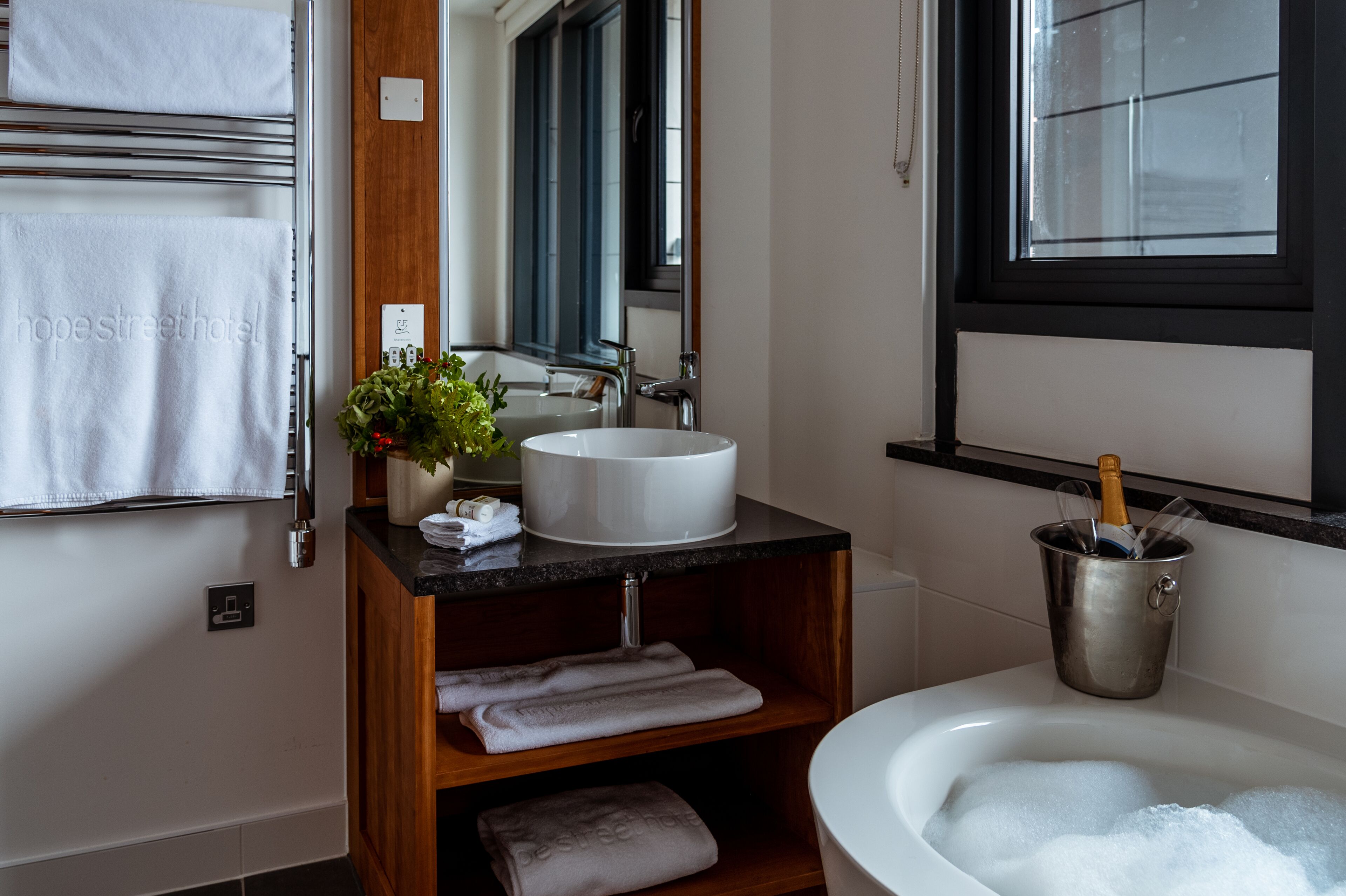 Un baño moderno con un lavamanos blanco, un espejo grande, un mueble de tocador de madera, una bañera y un cubo para champán.