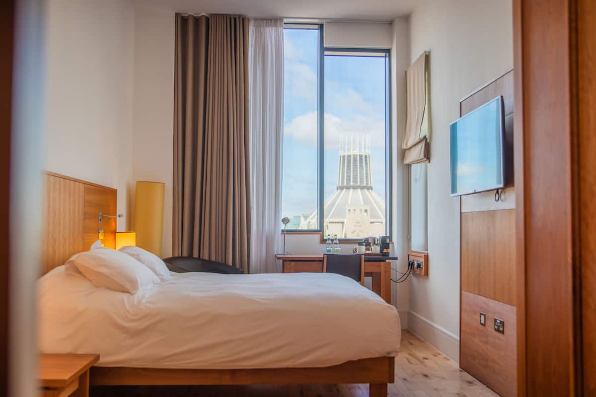 A hotel room with a bed, a desk, a chair, a TV, and a window with a cityscape view.