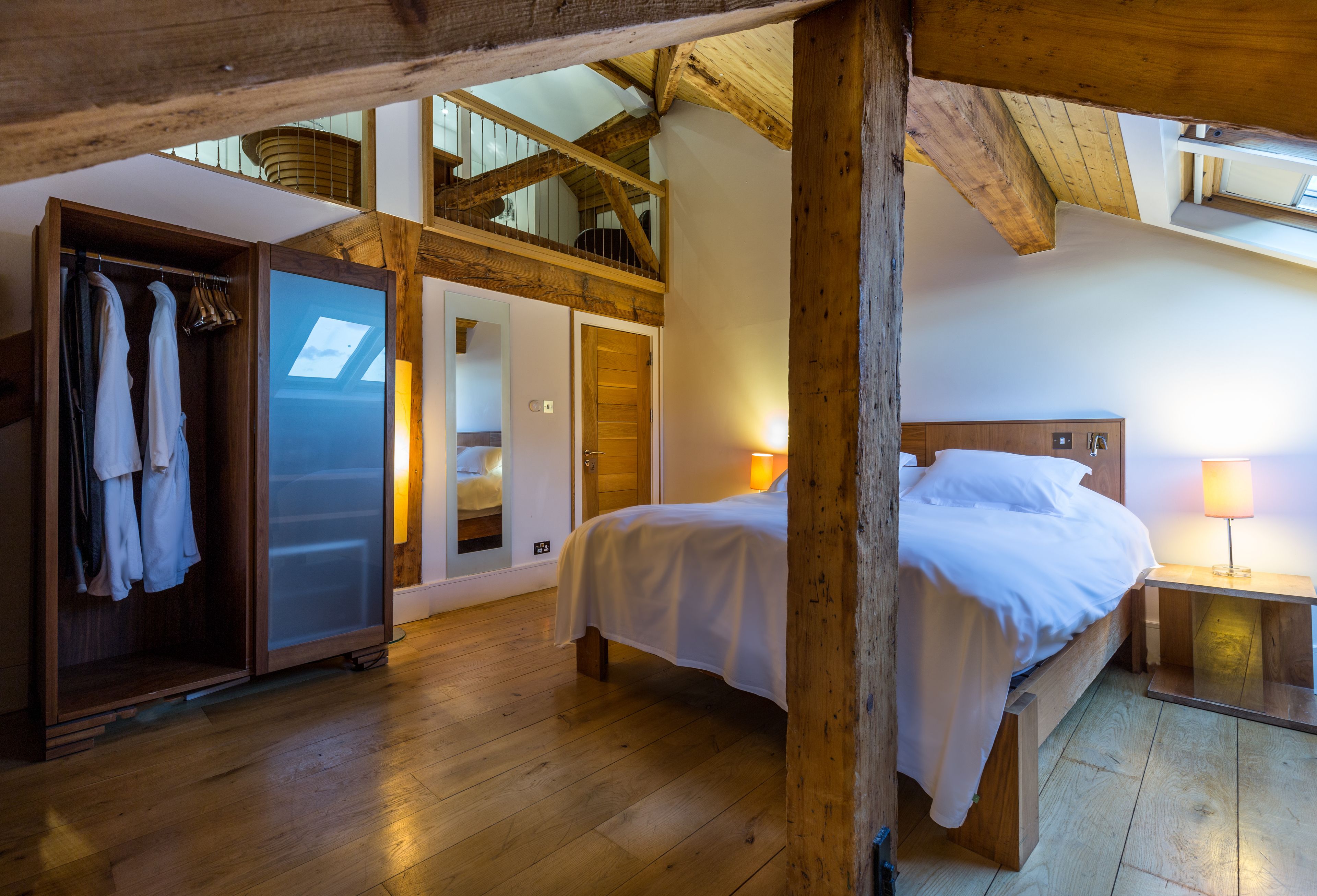 Un dormitorio con techo de madera, una cama con ropa de cama blanca, una mesita de noche con lámpara y un armario con ropa colgada.