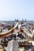 Two people toasting with champagne glasses, overlooking a cityscape with buildings and a clear sky.