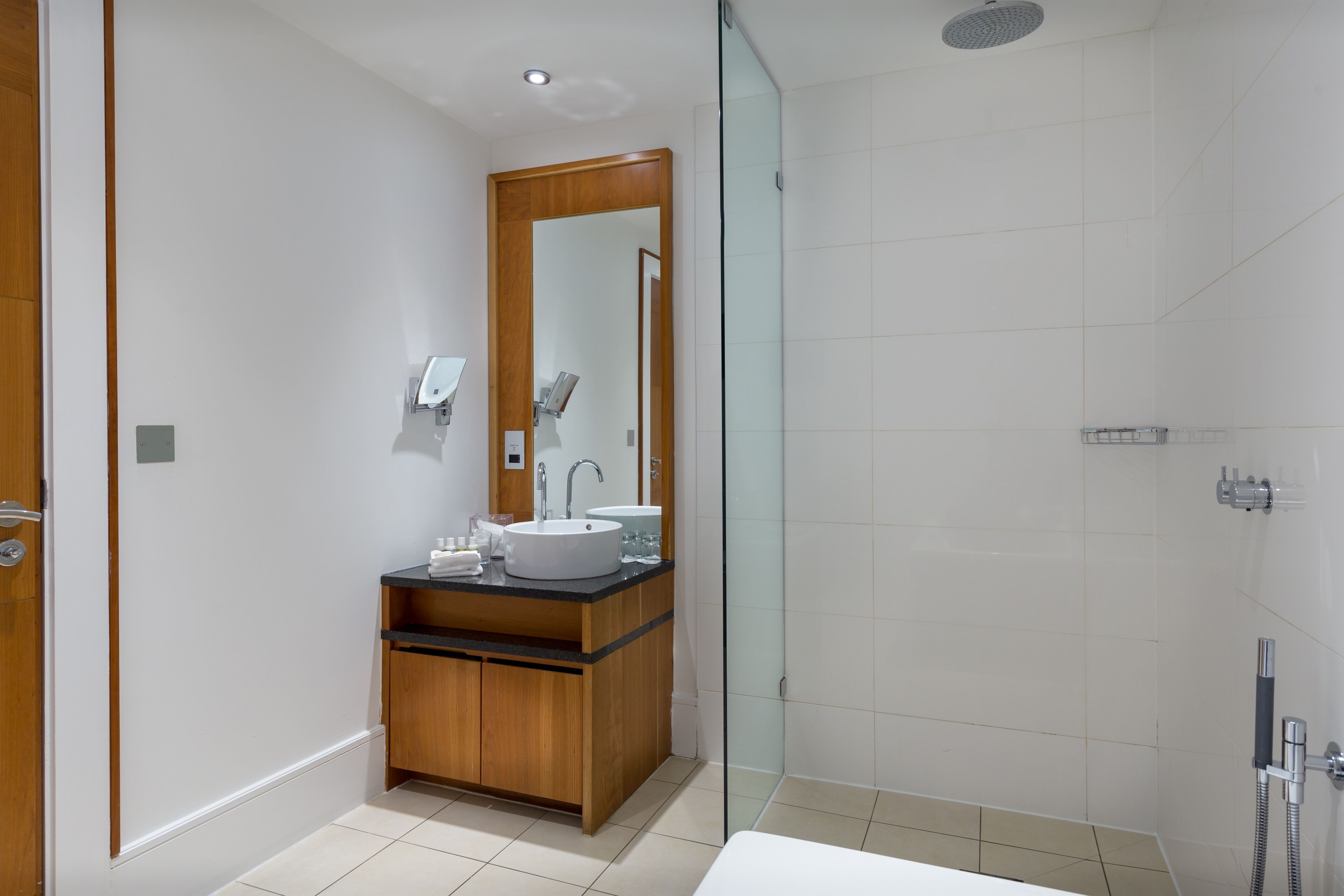Un baño moderno con un tocador de madera, un espejo grande, un lavamanos blanco y una zona de ducha con mampara de vidrio.