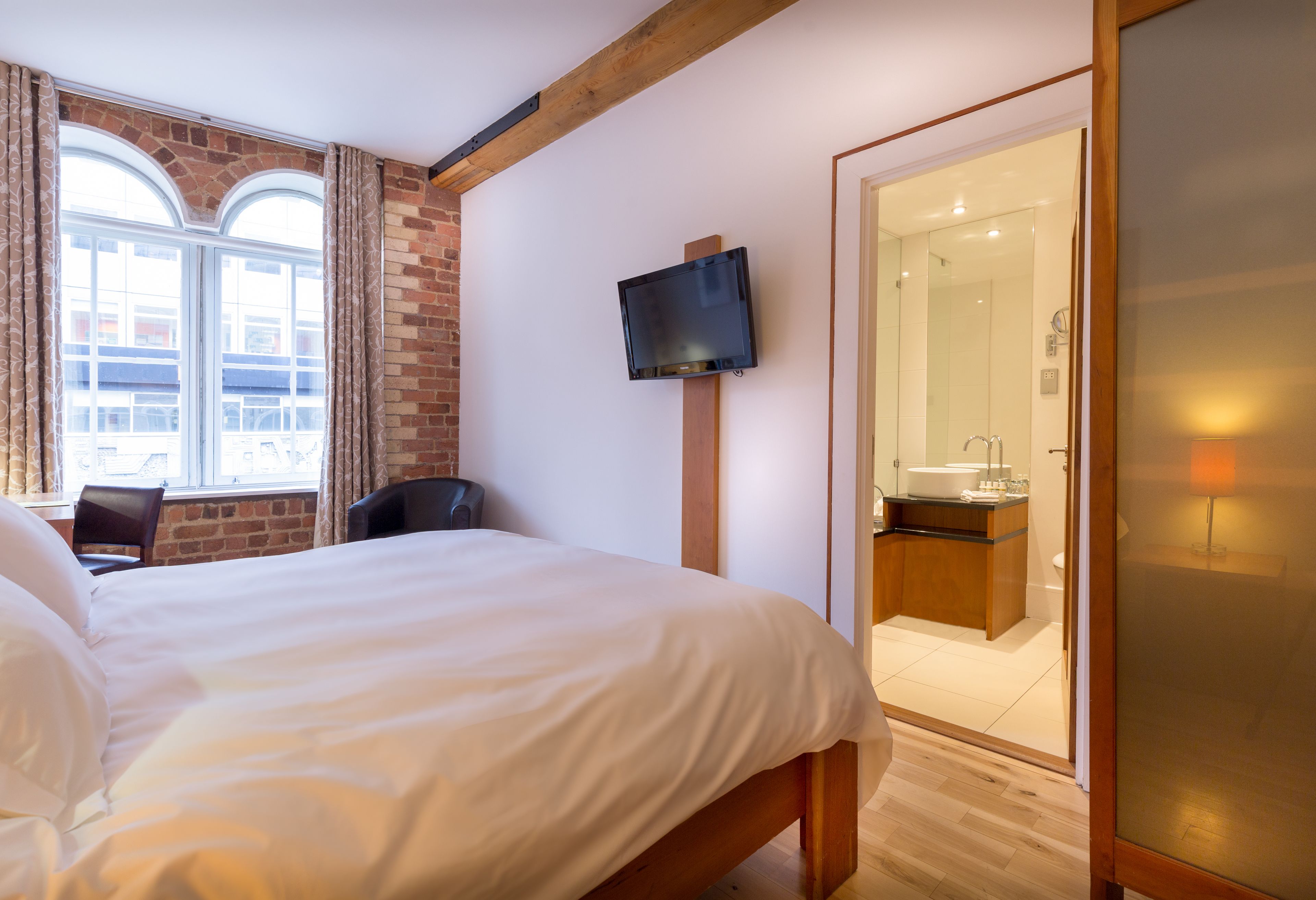 A hotel room with a large bed, a TV mounted on the wall, a wooden chair, and a view of the outside through a window with curtains.