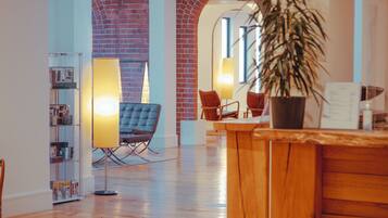 A spacious lobby with a wooden reception desk, a seating area with chairs, a bookshelf, and a large potted plant.