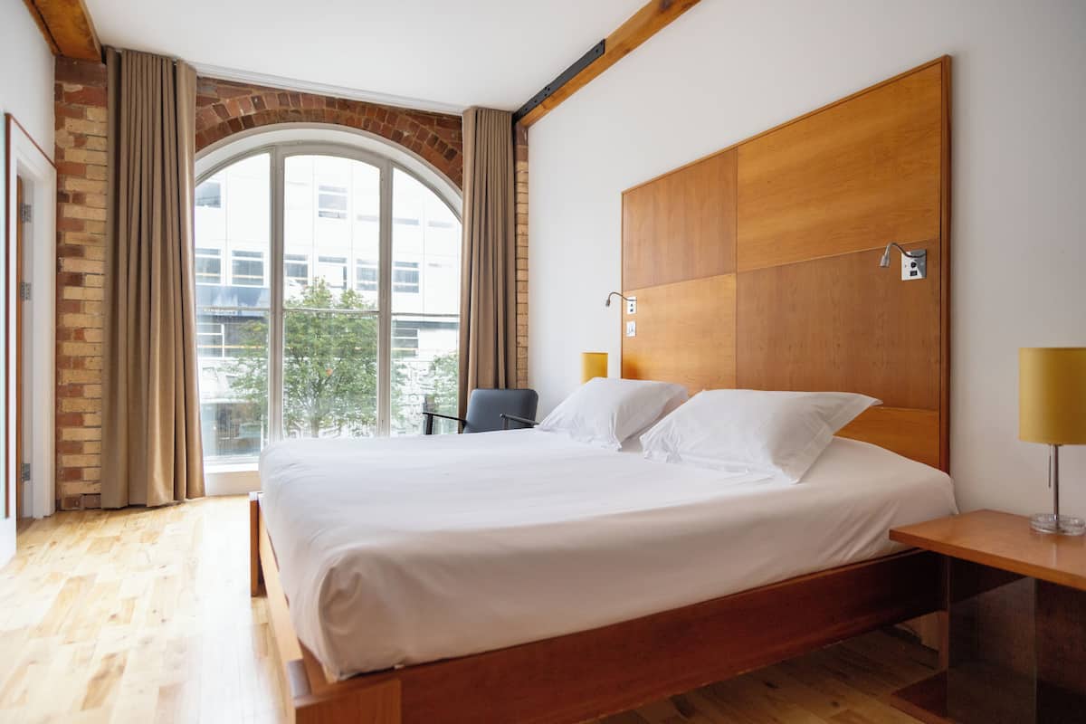 A hotel room with a large bed, wooden headboard, and a view of the city through a large window.