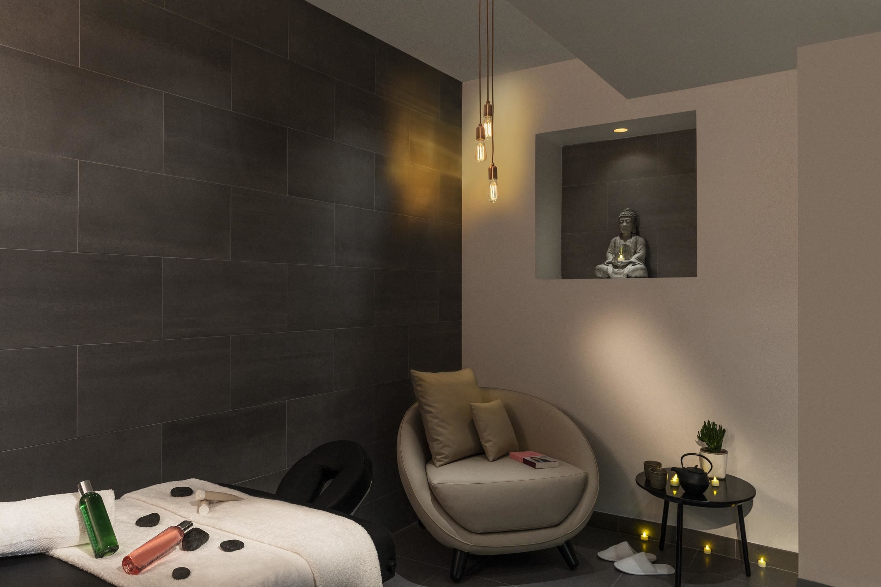 Une salle de spa moderne avec un mur recouvert de carreaux sombres, un fauteuil rond, une petite table avec des bougies et une statue représentant une figure en méditation.
