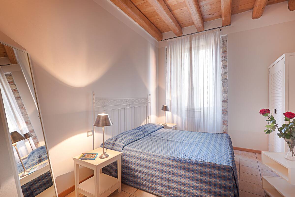 A bedroom with a bed, bedside tables, a mirror, a window with curtains, and a dresser with a vase of flowers.