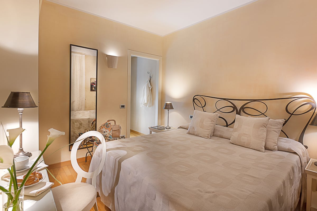 A bedroom with a large bed, a chair, a table with a lamp, and a mirror.