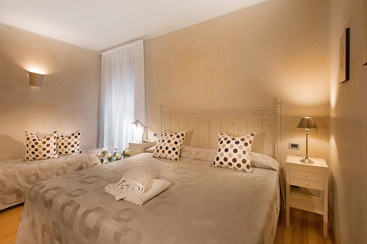 A hotel room with two beds, a nightstand, a lamp, and a window with curtains.
