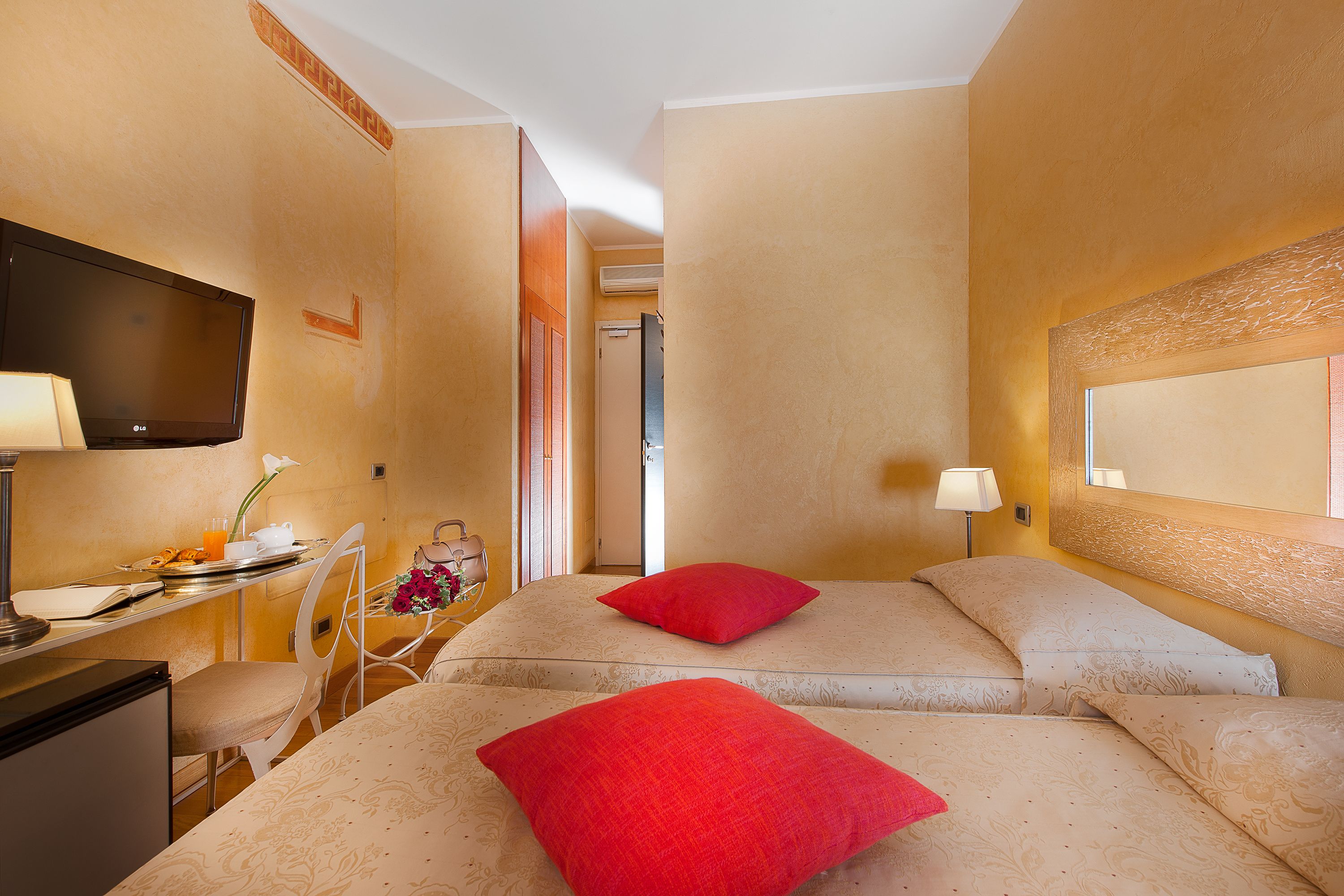 A hotel room with a bed, a desk with a television, a chair, and a bedside table with a lamp.