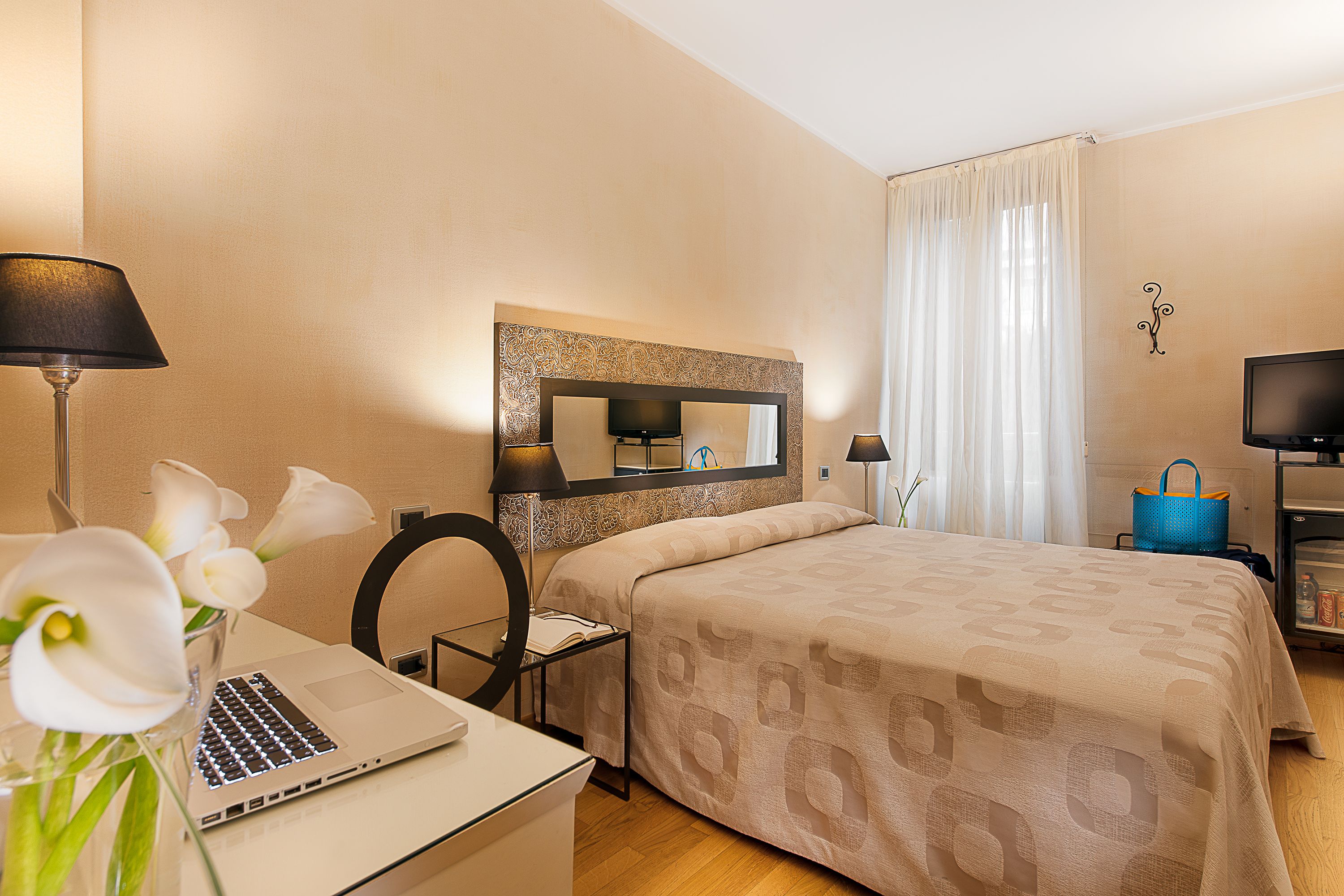 Habitación de hotel con cama, escritorio, computadora portátil, espejo, televisor y un florero con flores.
