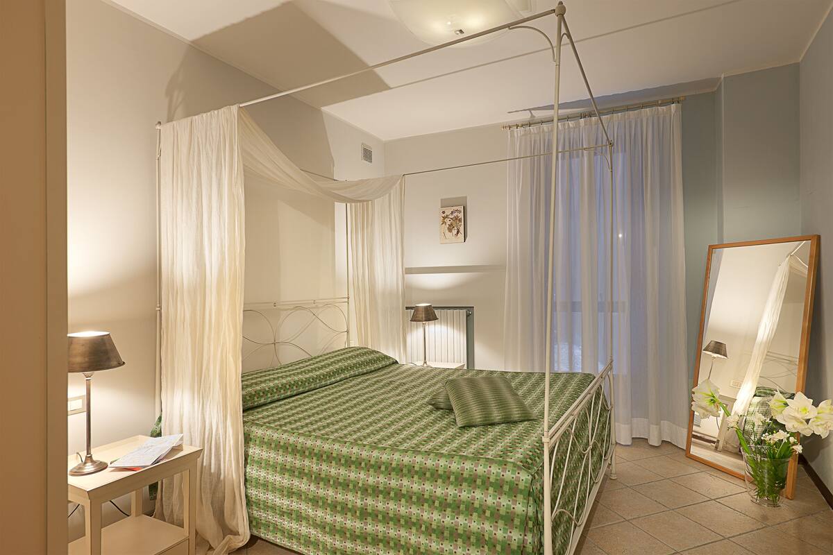 A bedroom with a four-poster bed, a bedside table with a lamp, a mirror, and a window with curtains.
