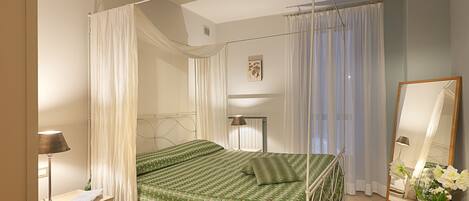 Un dormitorio con cama con dosel, una mesita de noche con lámpara, un espejo y una ventana con cortinas.