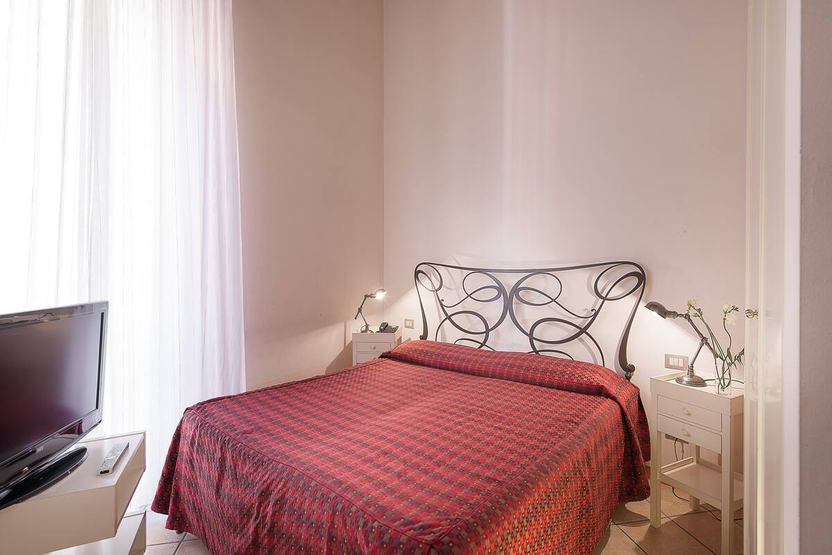 A neatly made bed with a patterned bedspread, a bedside table with a lamp, and a TV on a stand.