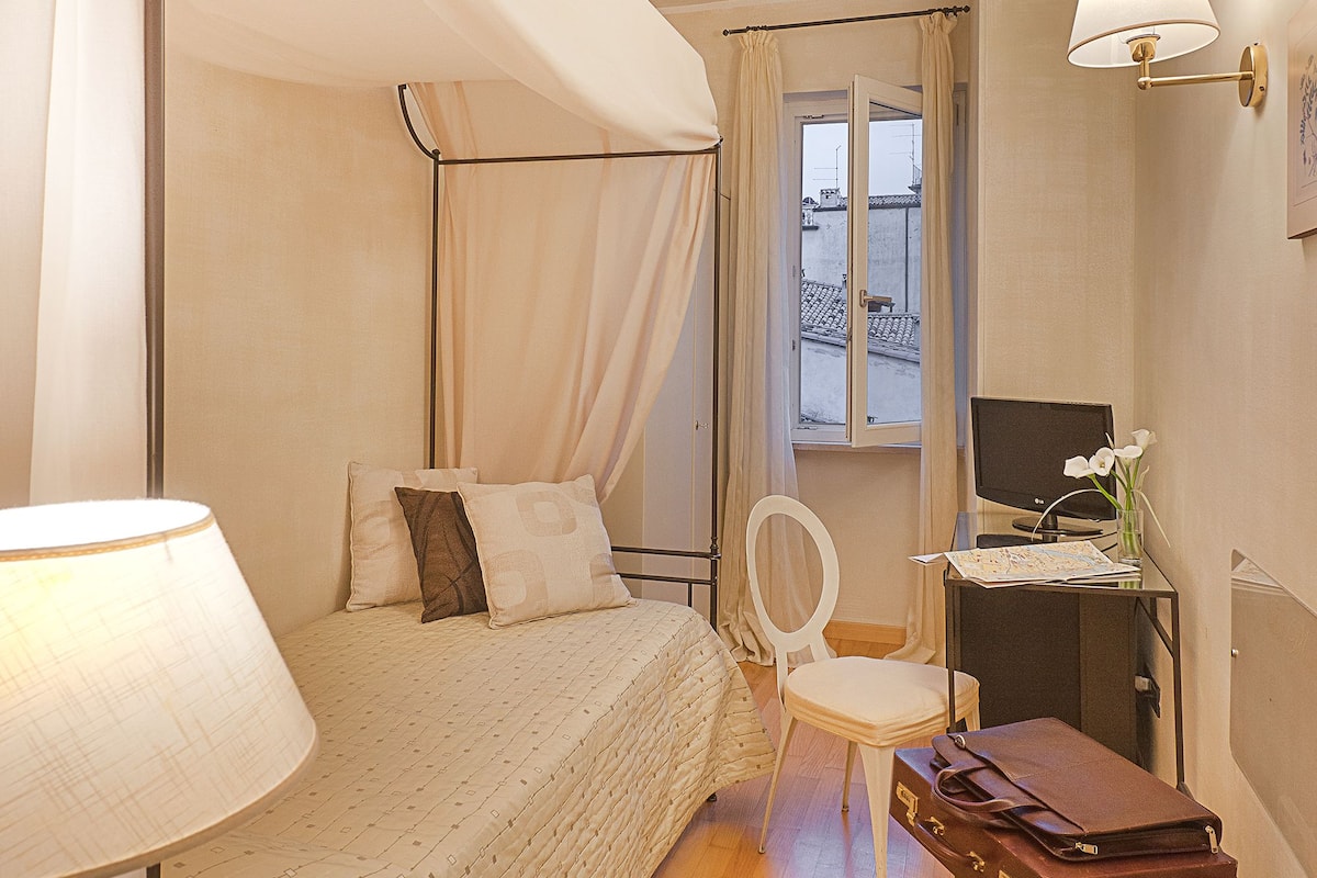 A bedroom with a canopy bed, a desk with a lamp, a chair, and a suitcase.