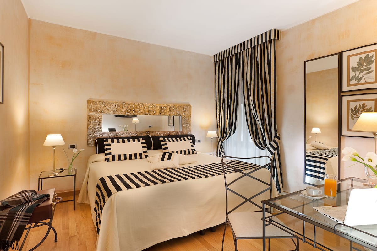 A hotel room with a bed, bedside tables, a glass table, a chair, a mirror, and curtains.