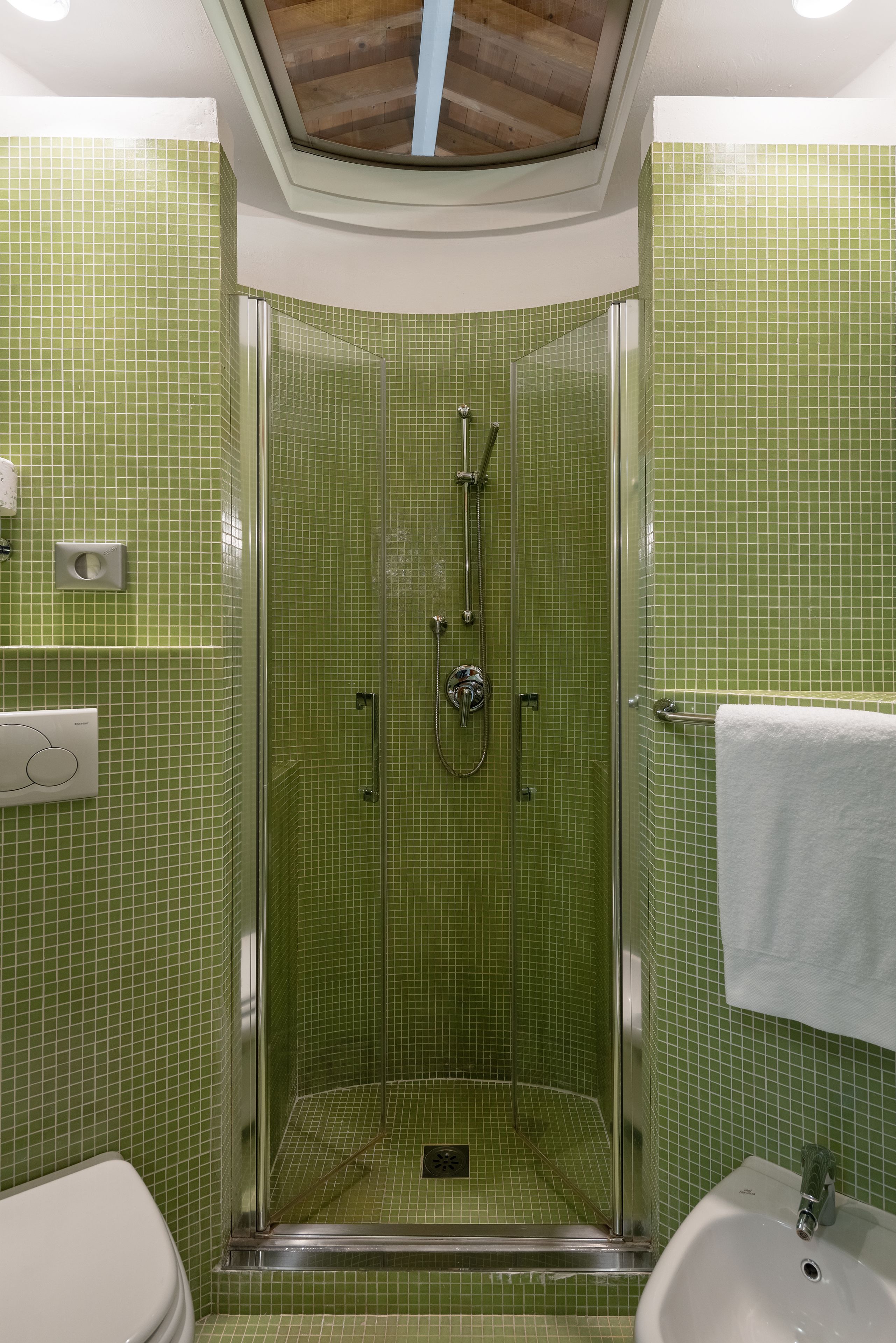 Un baño con paredes de azulejos verdes, una ducha con mampara de vidrio, un inodoro y un lavamanos con una toalla colgada en la pared.