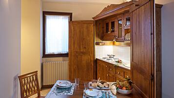 A wooden dining table with chairs, a window with curtains, and a kitchen area with wooden cabinets and a tiled floor.