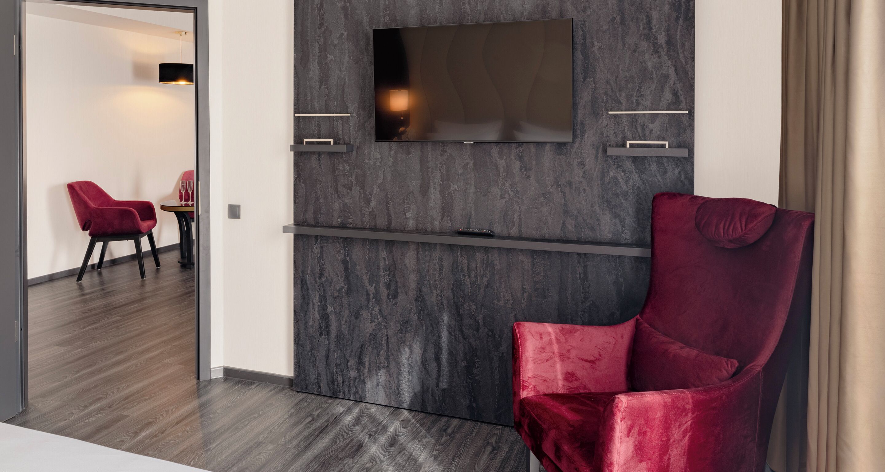 Una sala de estar moderna con un televisor de pantalla plana montado en una pared texturizada, un sillón de terciopelo rojo y un piso de madera oscura.