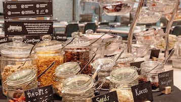 Ein Buffet mit verschiedenen Speisen, darunter 'Cakes', 'Cornflakes', 'Choco pops', 'Rosinen', 'Kokos' und 'Samen'.